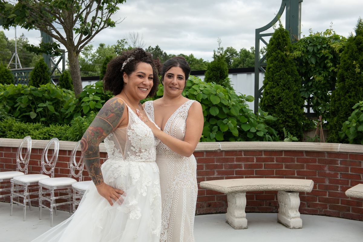Two brides in lace gowns stand smiling in a garden courtyard with lush greenery and a stone bench behind them.