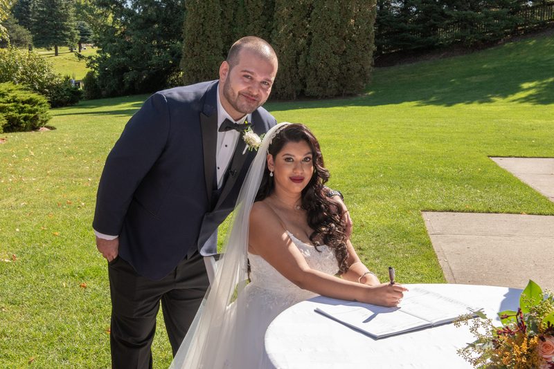 A bride signs documents at a white-clothed table with her smiling groom beside her in a sunlit outdoor setting.