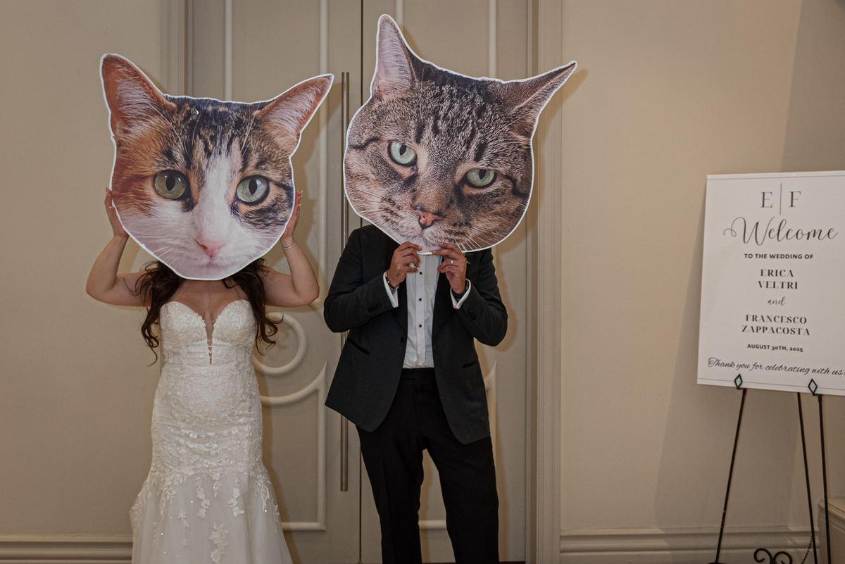 A bride and groom hold large cat-face cutouts over their own faces beside a welcome sign in a warmly lit entrance.