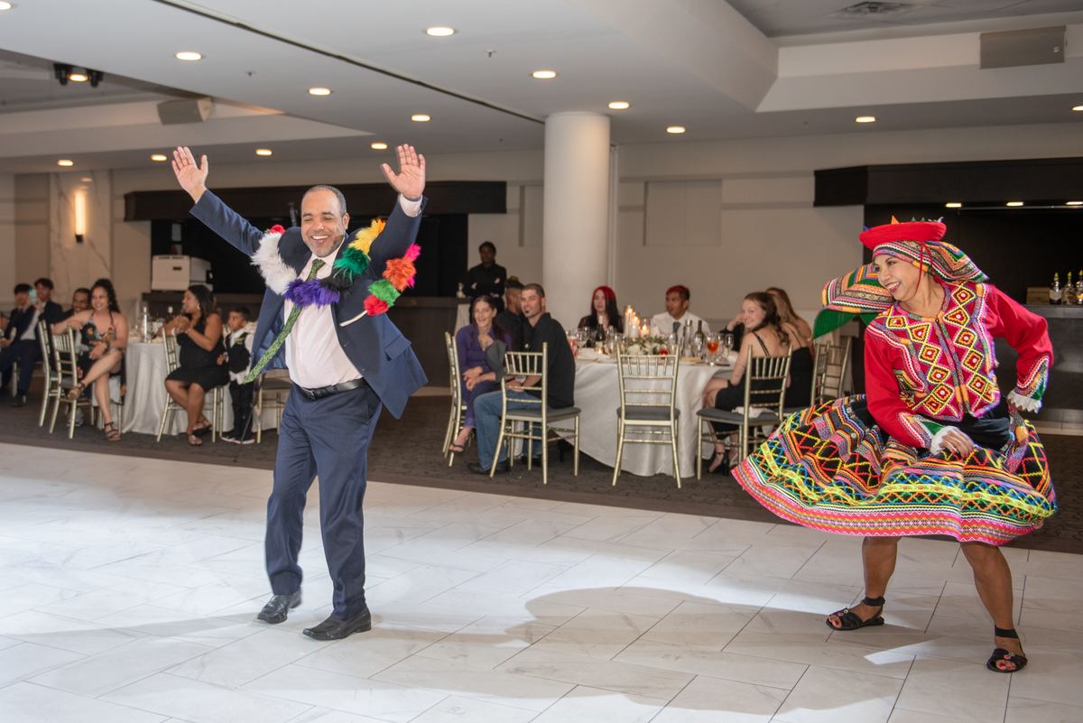 A man in a suit with feathers and a woman in folk attire dance joyfully at a banquet hall, celebrating.