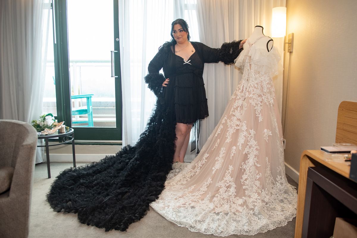 A woman in a black feathered gown stands next to a mannequin displaying a white lace wedding dress in a well-lit room.