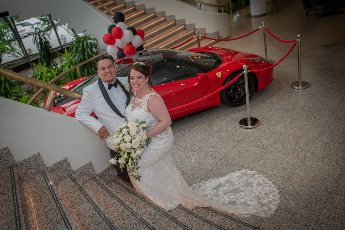 A bride in a lace gown and a groom in a tuxedo smile on a grand staircase beside a red Ferrari and clusters of balloons.