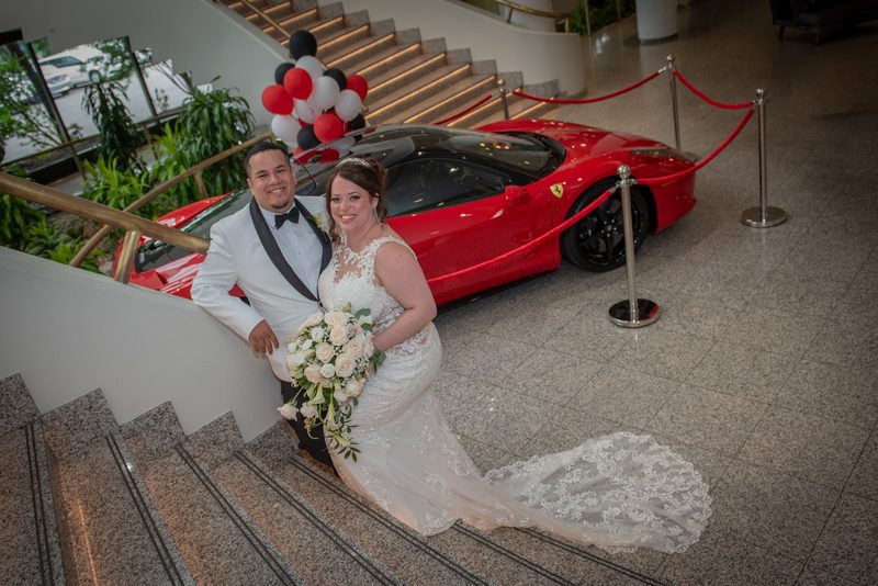 A bride in a lace gown and a groom in a tuxedo smile on a grand staircase beside a red Ferrari and clusters of balloons.