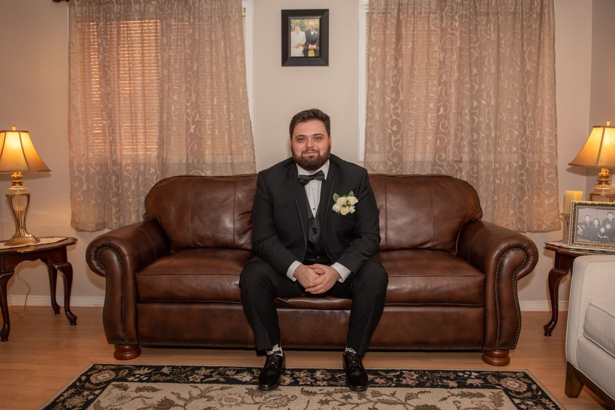 A groom in a black tuxedo smiles at the camera on a leather sofa in a warmly lit living room with family photos behind him.