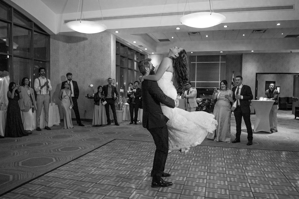 A bride and groom leap mid-air in a black-and-white reception dance while guests smile and raise their phones to catch it.