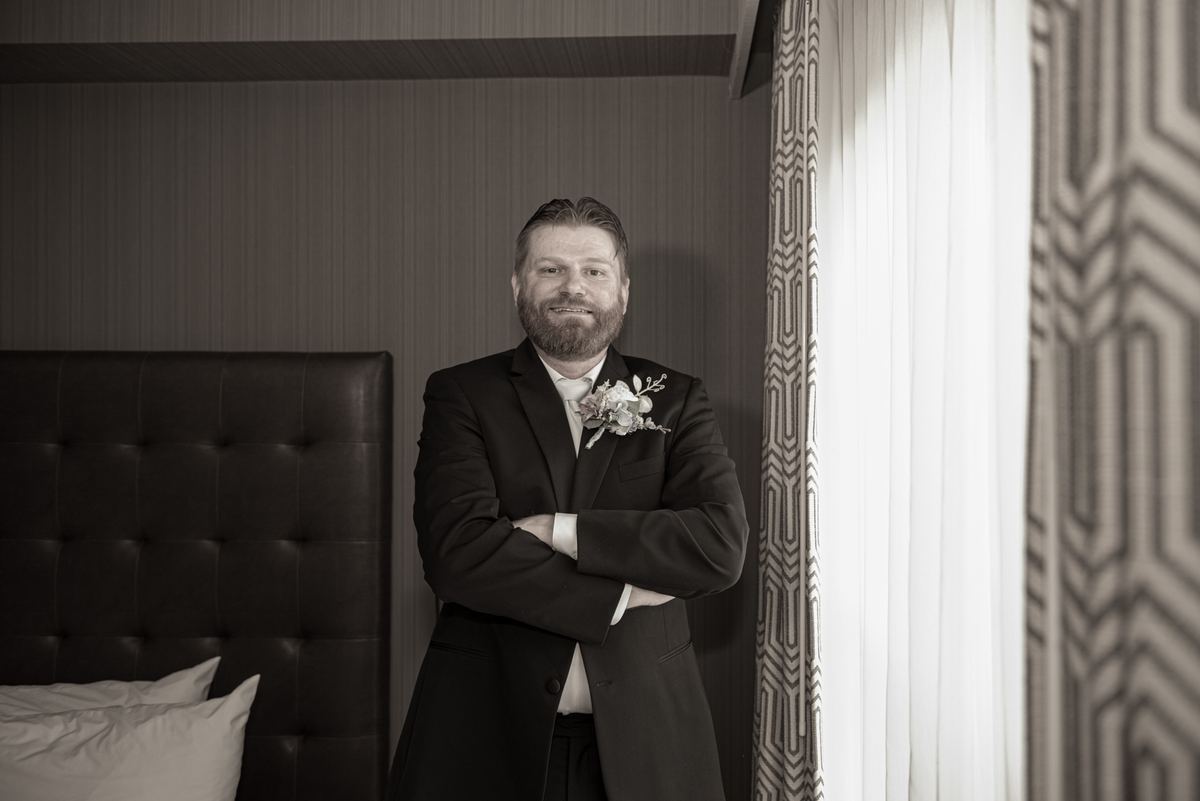 A groom stands confidently in a hotel room, smiling at the camera with his suit jacket and boutonnière visible.
