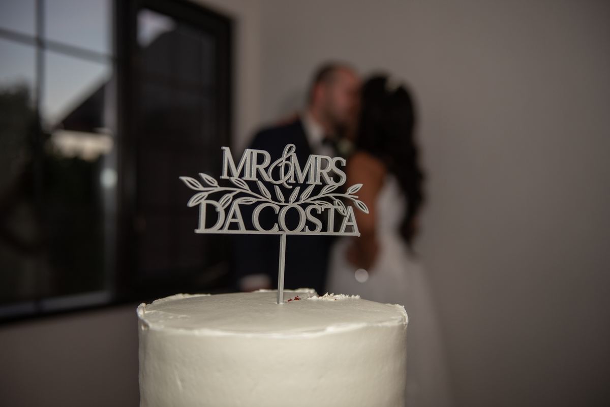 A bride and groom kiss in the background while their wedding cake with a Mr & Mrs topper is in sharp focus.