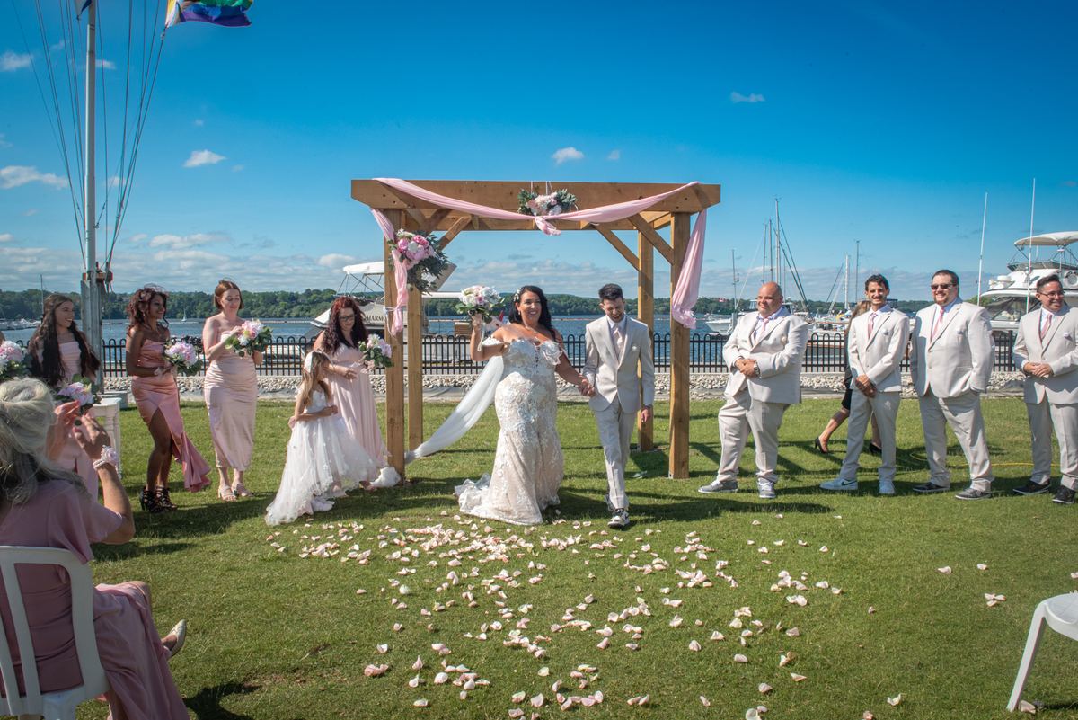 Bride and groom walk down grassy aisle toward wooden arch adorned with pink ribbons, surrounded by wedding party.