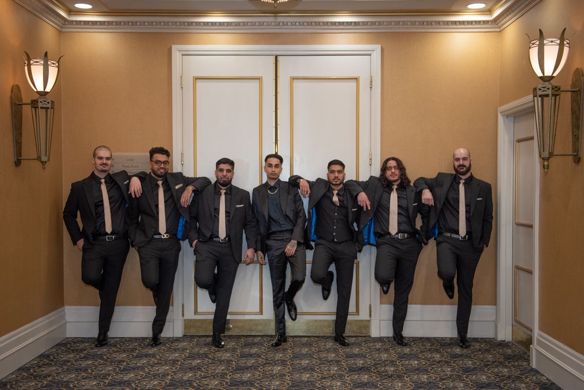 Seven men in black suits pose playfully in a grand hallway with gold-trimmed doors and patterned carpet.