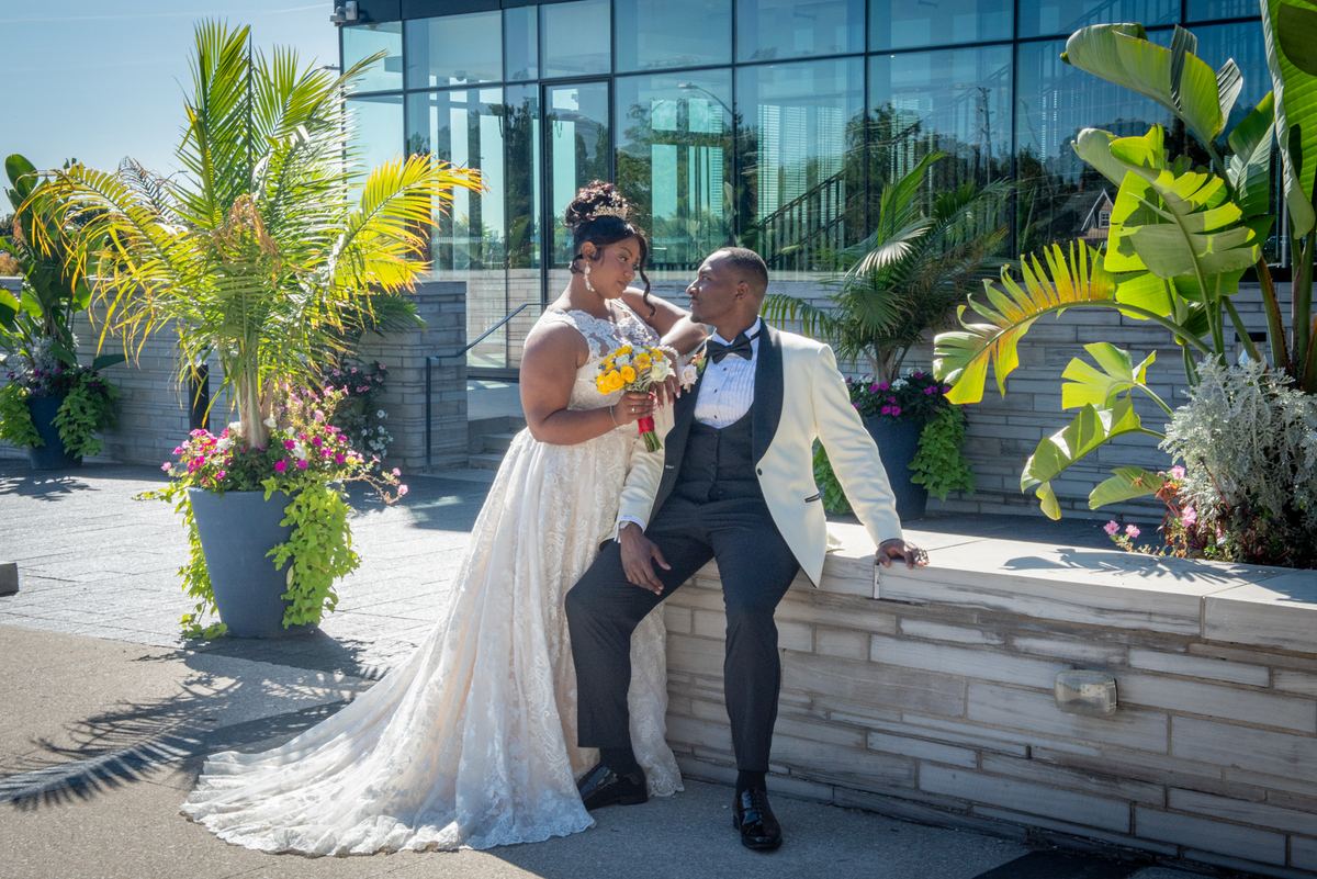 A bride in white touches her groom's face as they sit by a stone ledge surrounded by tropical foliage.