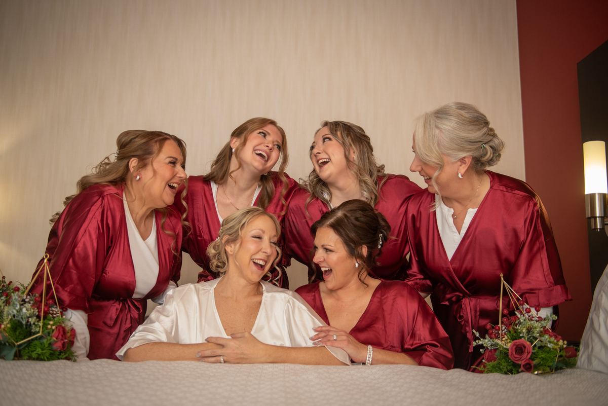 Bridesmaids in burgundy satin surround the bride in white, laughing around a bed with floral arrangements.