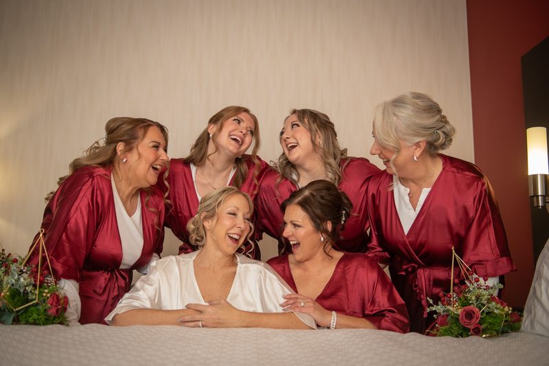 Bridesmaids in burgundy satin surround the bride in white, laughing around a bed with floral arrangements.