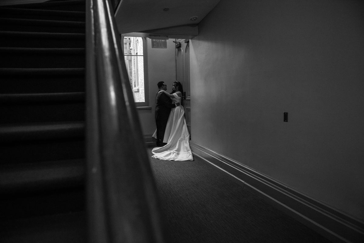 A bride and groom embrace tenderly in a dim hallway, illuminated by soft light from an adjacent window.