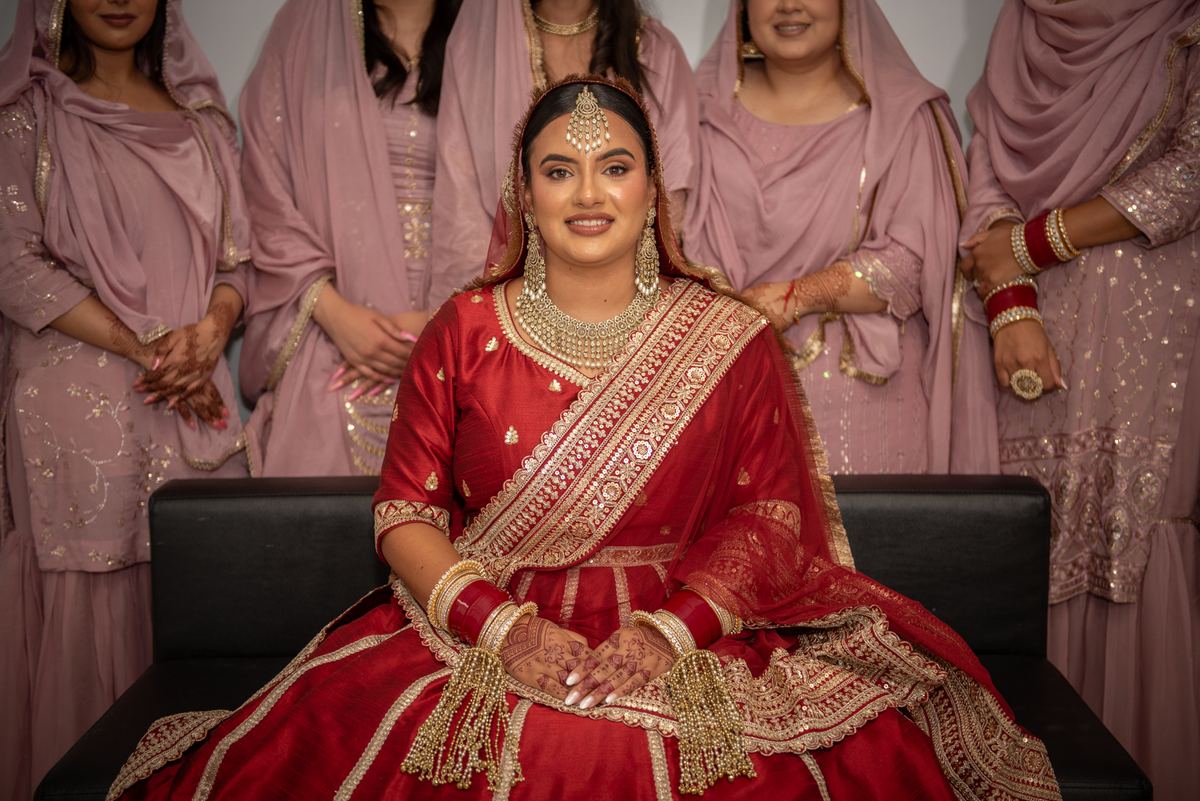 A bride in a red and gold lehenga sits on a black couch, smiling at the camera with bridesmaids behind her.
