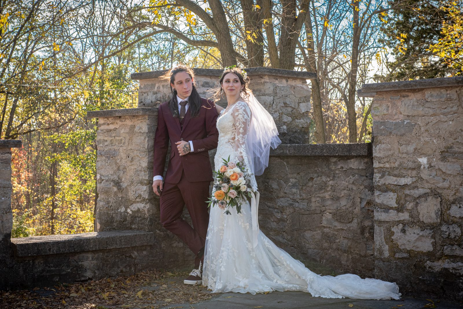 Bride in white lace gown and veil beside groom in burgundy suit near stone wall under autumn sunlight.