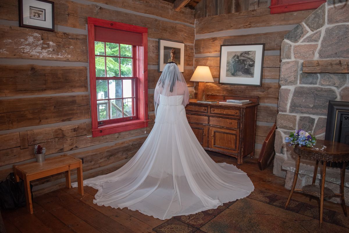 A bride in a white gown stands by a red-framed window in a rustic cabin, illuminated by soft light from a lamp.