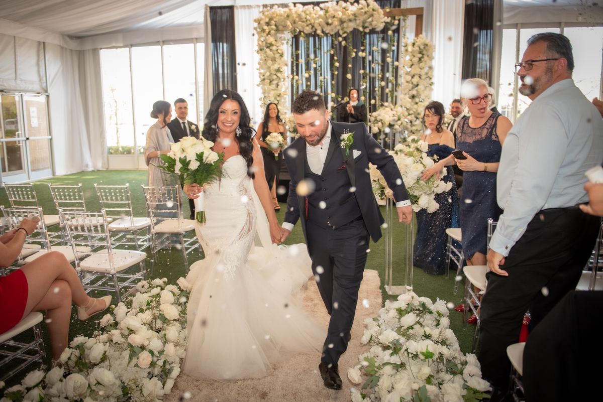 Bride and groom walk down the aisle under a tent filled with guests and flowers, confetti falling around them.