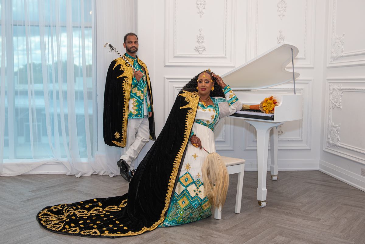 A man and woman in ceremonial attire stand beside a grand piano in an elegant room, exuding regal presence.