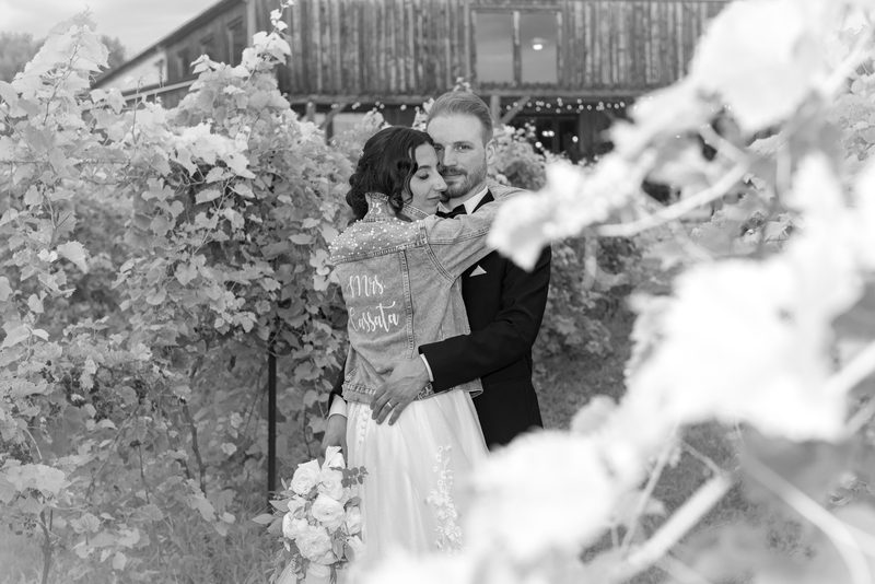 Bride in a denim jacket embroidered with "Mrs. Cassata" and groom embrace in a vineyard, surrounded by out-of-focus foliage.