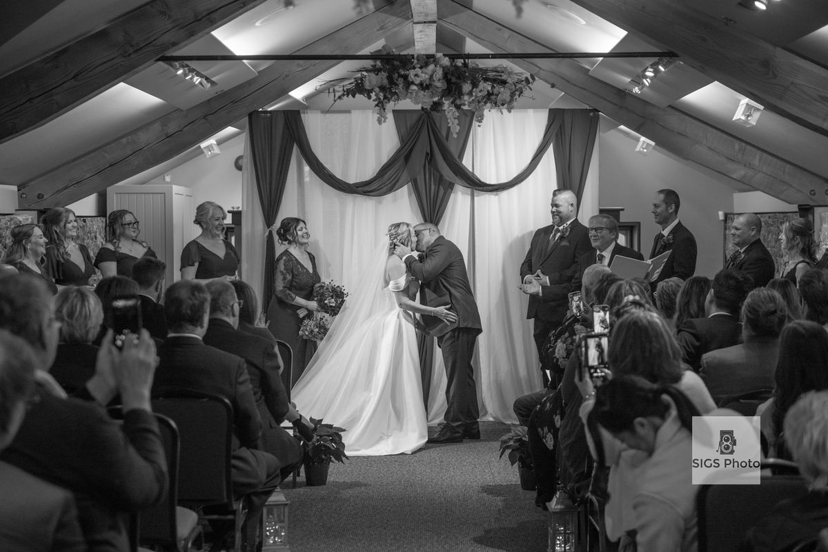 A bride and groom kiss at the altar, surrounded by smiling guests in a warmly lit space with floral decorations.