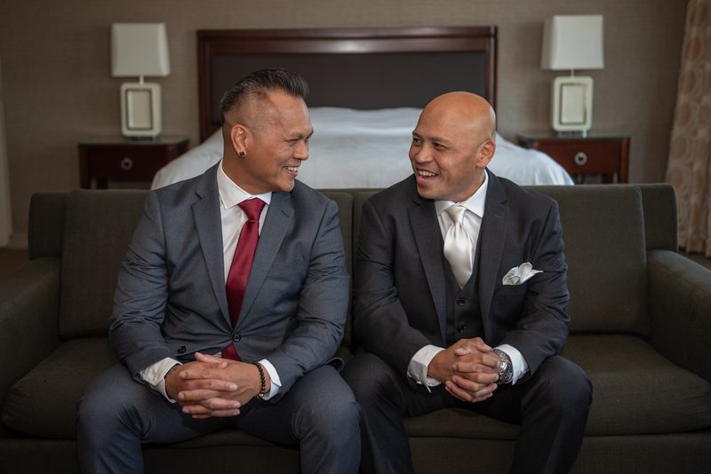 Two men in formal suits sit on a couch, laughing warmly under soft bedside lamp lighting in a hotel room, celebrating.