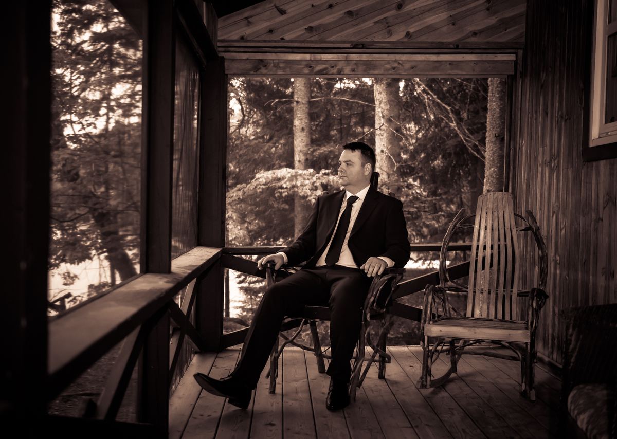 A man in a suit sits thoughtfully on a wooden porch, looking out at a forest illuminated by golden sunlight.