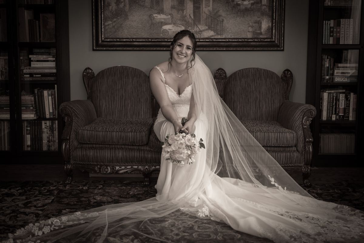 A smiling bride in an elegant gown sits on a sofa, holding her bouquet and surrounded by vintage decor.