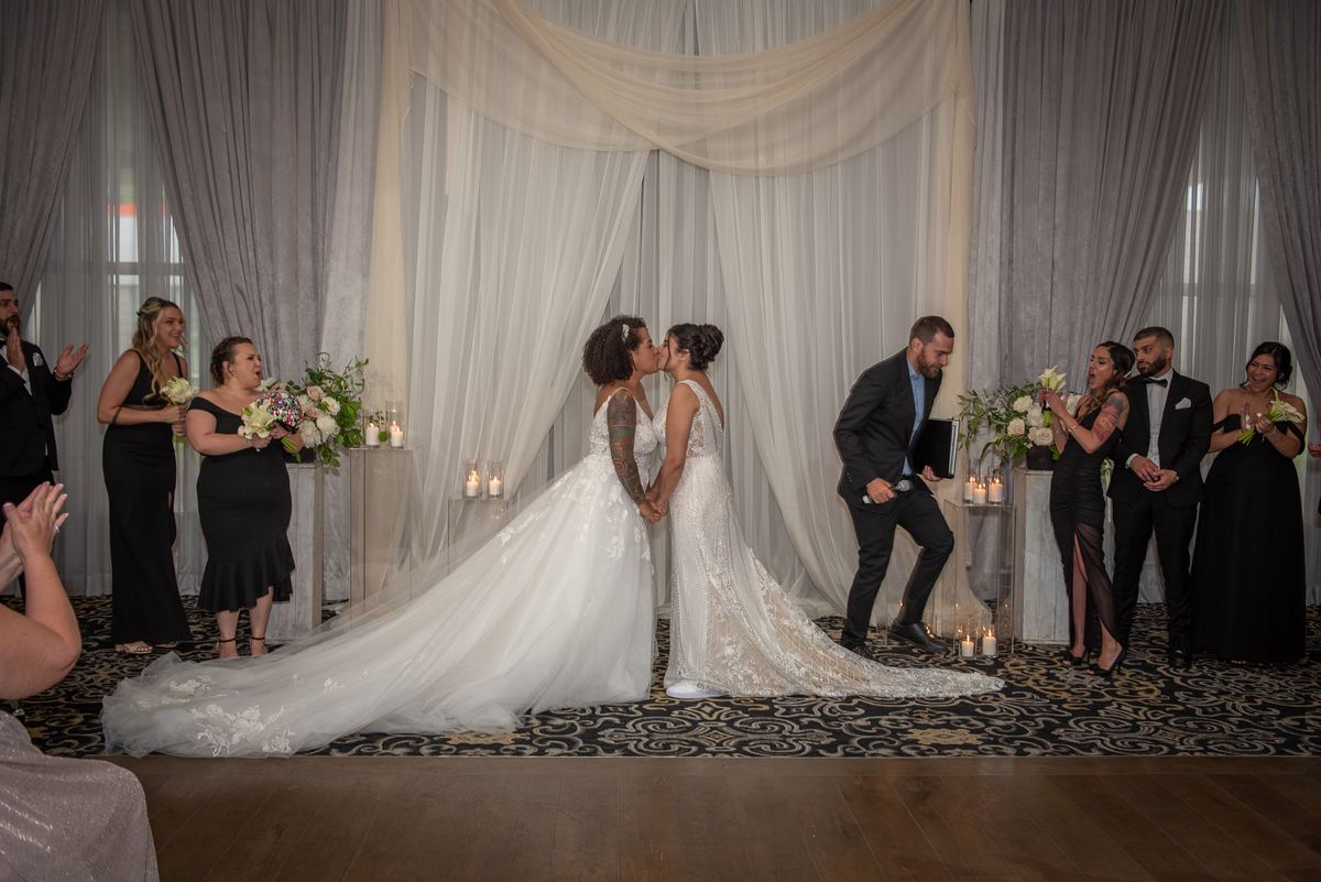 Two brides kiss at the center of an elegant draped backdrop, surrounded by guests in formal attire and bouquets.