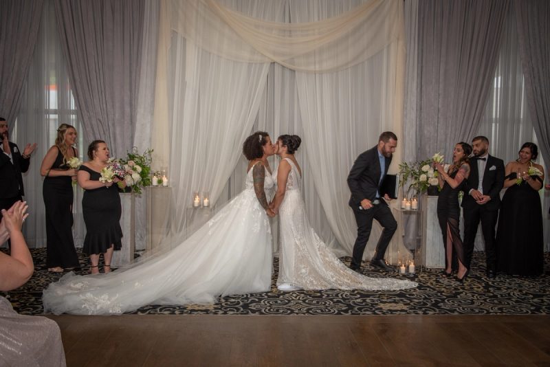 Two brides kiss at the center of an elegant draped backdrop, surrounded by guests in formal attire and bouquets.
