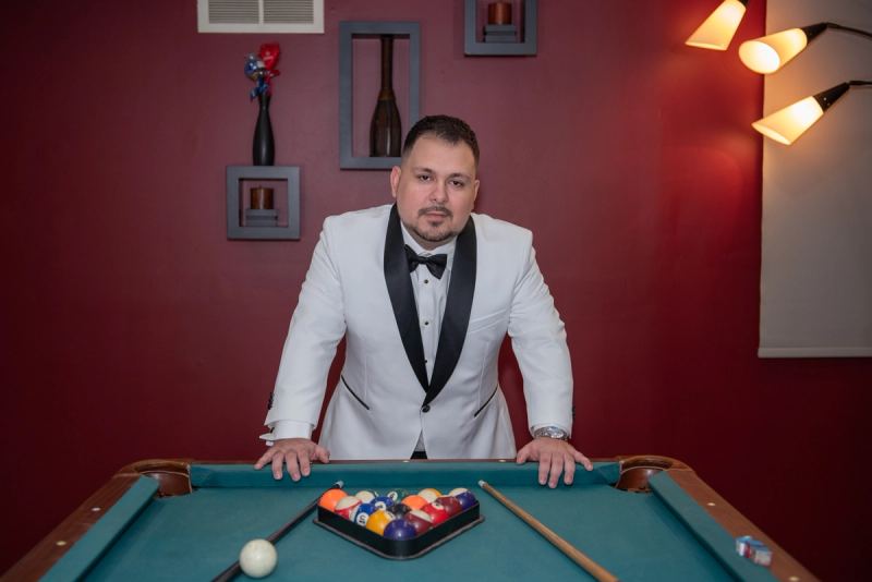 A man in a white tuxedo stands behind a pool table, hands on the felt, illuminated by warm sconces against a red wall.