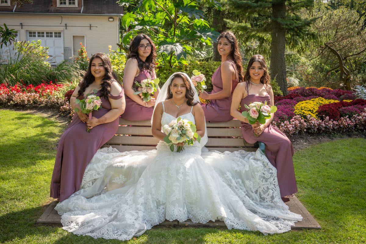 A bride and four bridesmaids in matching mauve dresses smile with bouquets of pink and white flowers.