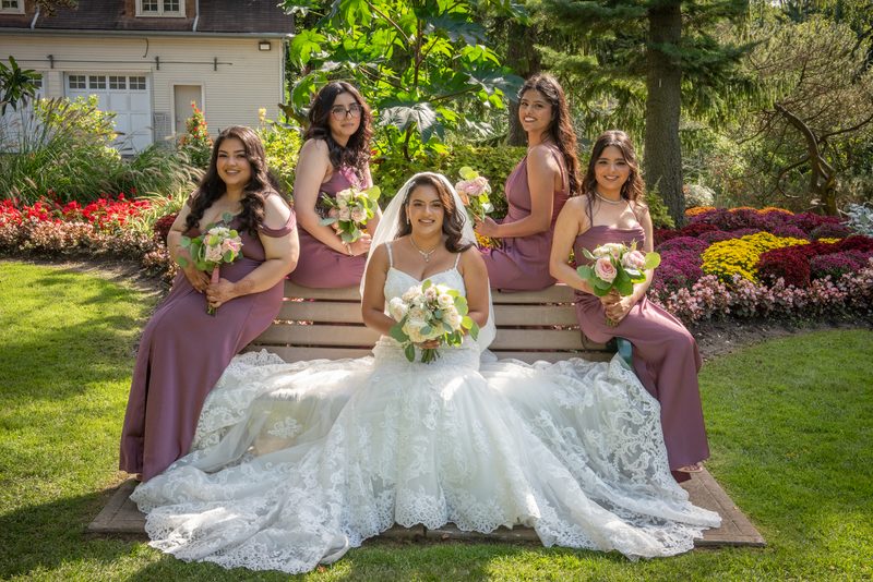 A bride and four bridesmaids in matching mauve dresses smile with bouquets of pink and white flowers.