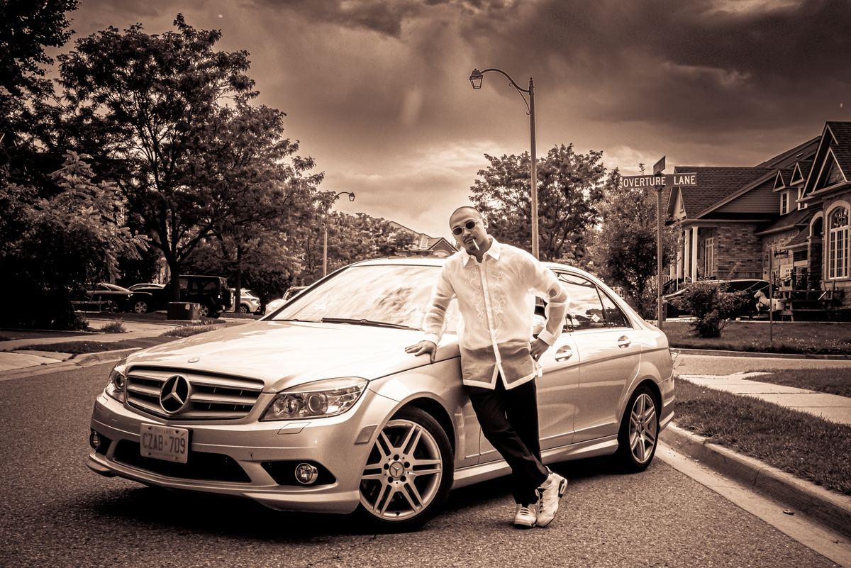 A man in a white shirt leans casually against a luxury Mercedes-Benz sedan on a suburban street under an overcast sky.