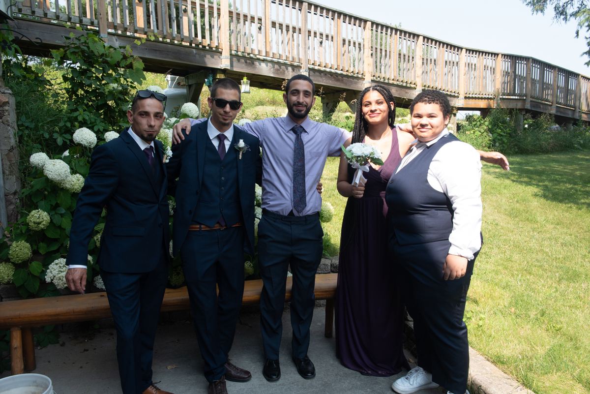 Five formally dressed people pose outdoors under a wooden bridge with lush greenery, one holding a small bouquet.