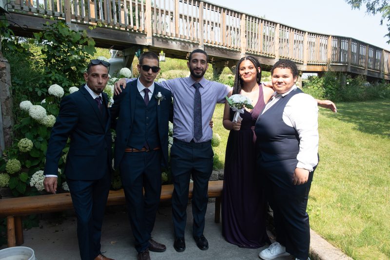 Five formally dressed people pose outdoors under a wooden bridge with lush greenery, one holding a small bouquet.