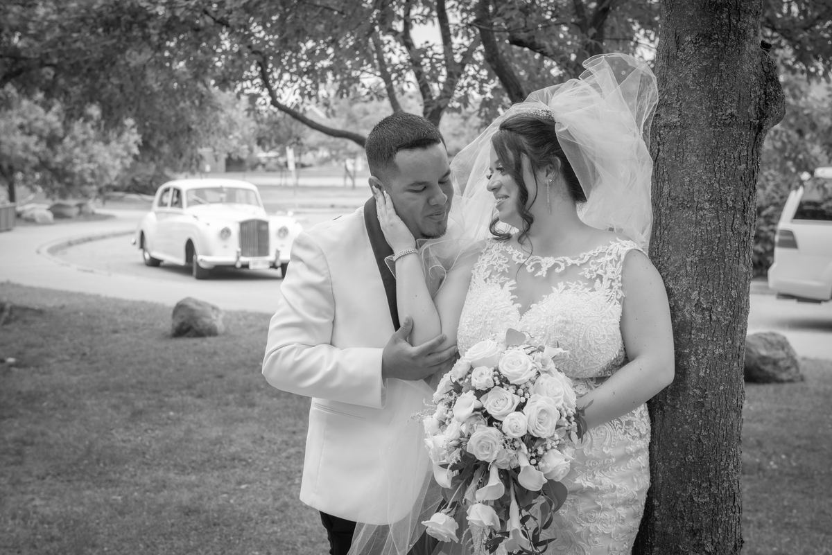 A newlywed couple shares an intimate moment outdoors, with the groom cradling the bride's face near a tree.