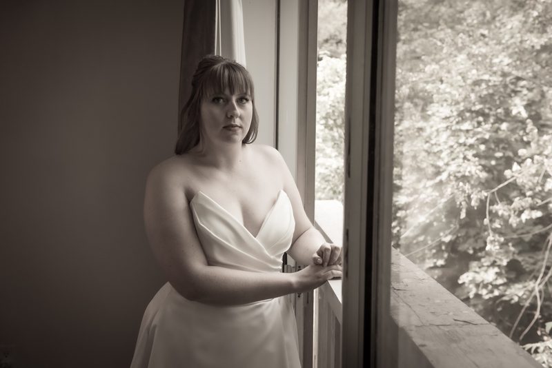 A bride in a strapless gown stands by an open window, hands resting on the sill, gazing into a sepia-toned.
