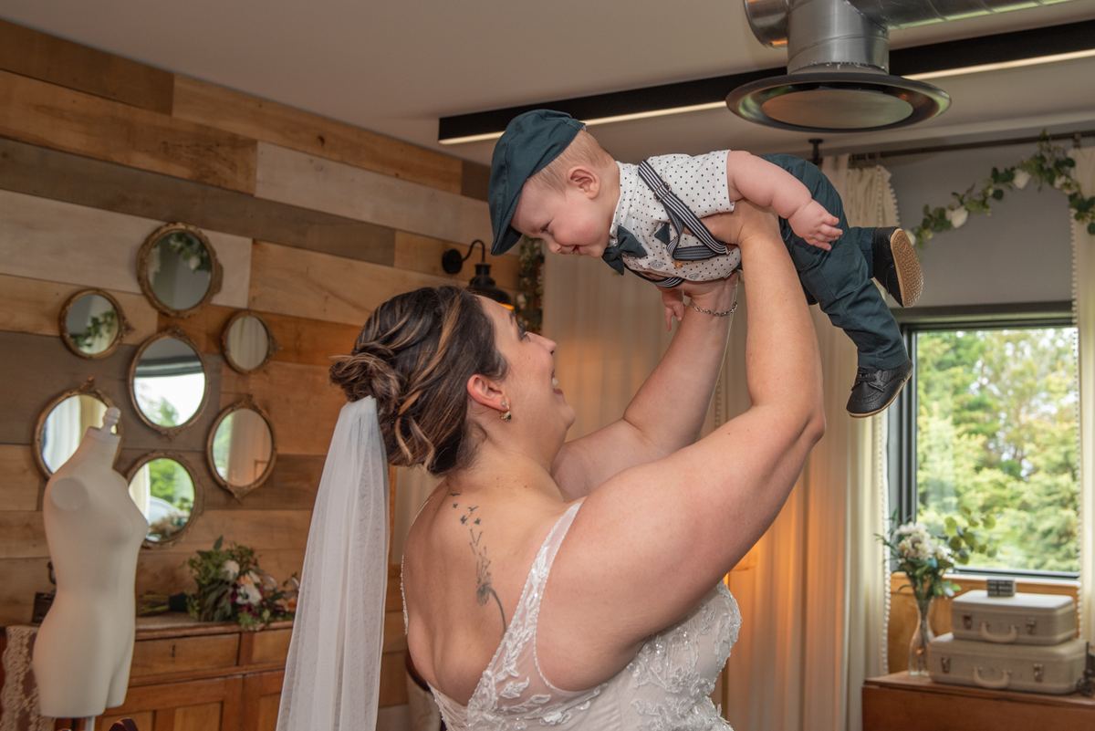 A bride holds her child high, both smiling joyfully in a rustic room with wooden walls and natural light.