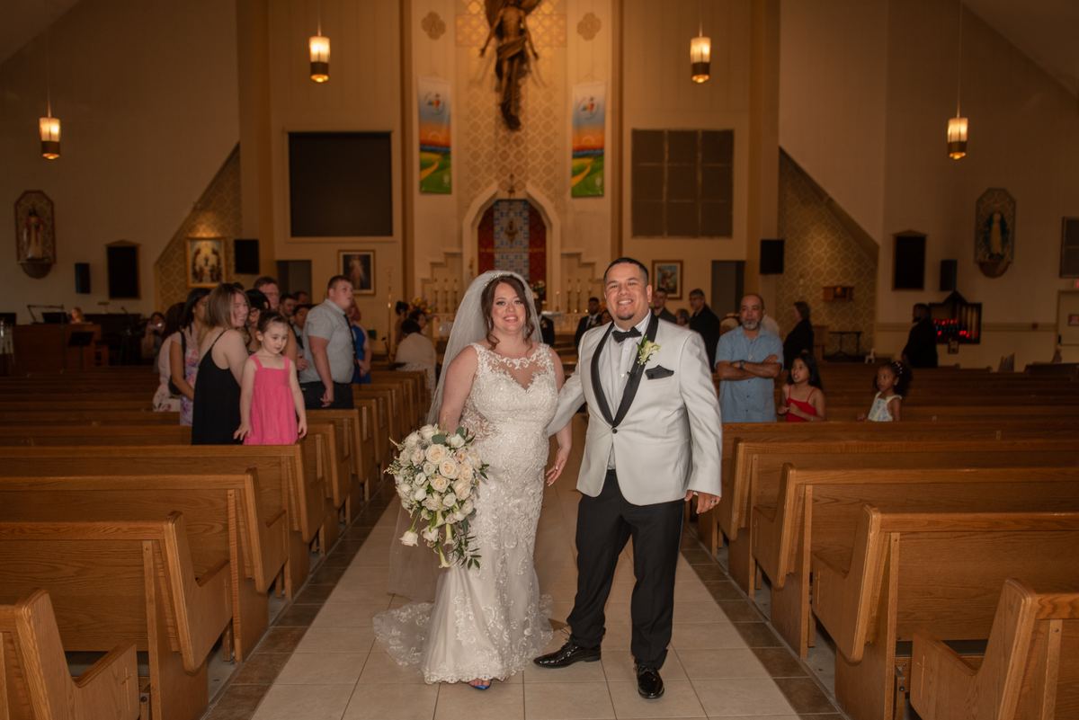 A smiling bride in a white dress and groom in a tuxedo exit a church aisle, guests watching from wooden pews.