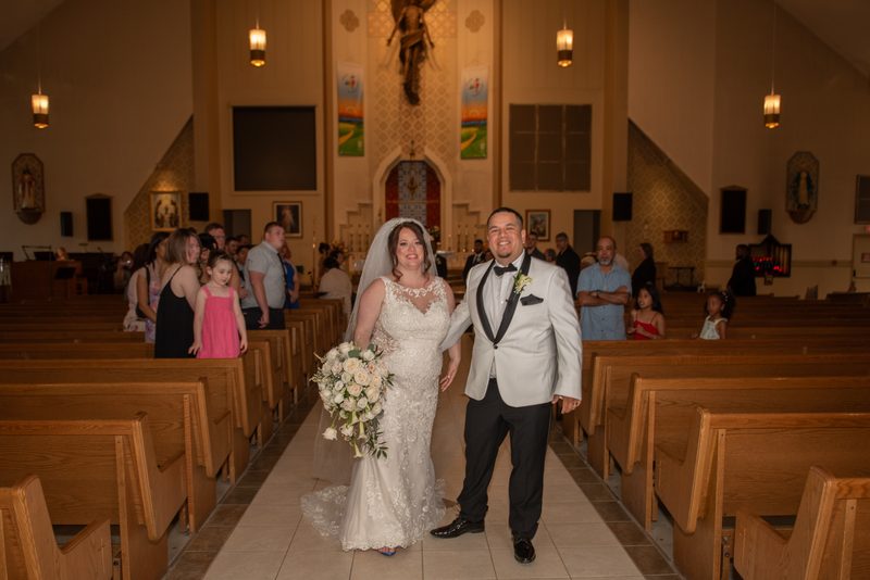 A smiling bride in a white dress and groom in a tuxedo exit a church aisle, guests watching from wooden pews.