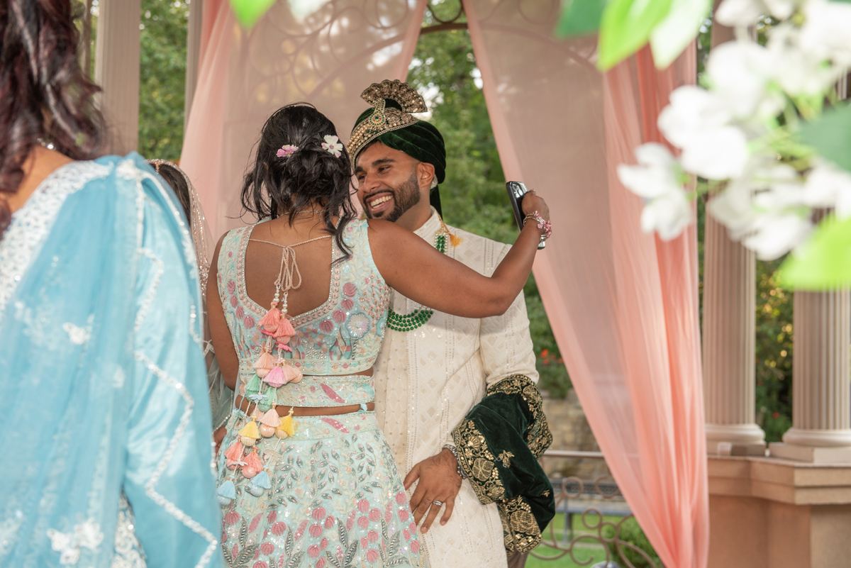 Bride and groom smiling under decorated canopy, she takes selfie with him, both in vibrant traditional attire.