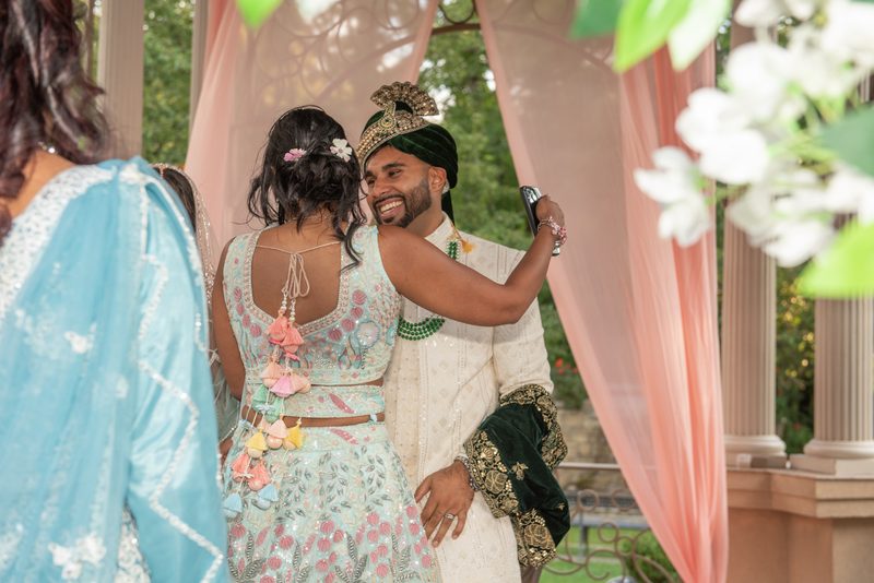 Bride and groom smiling under decorated canopy, she takes selfie with him, both in vibrant traditional attire.