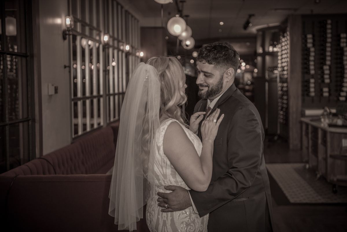 A bride and groom embrace tenderly, the bride adjusting the groom's lapel in a warmly lit room with modern decor.