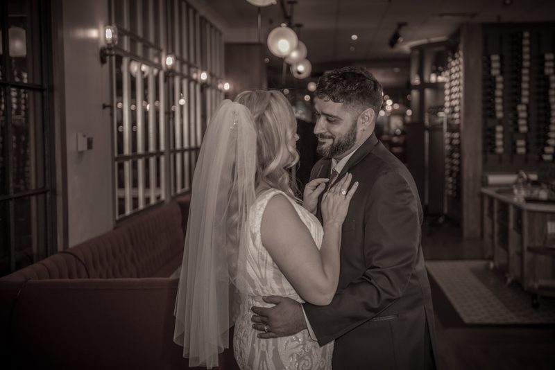 A bride and groom embrace tenderly, the bride adjusting the groom's lapel in a warmly lit room with modern decor.