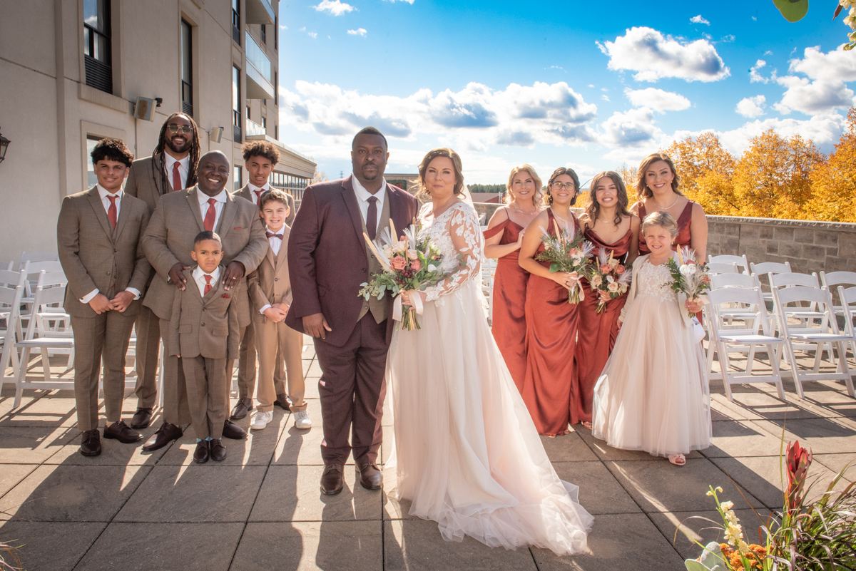 A bride and groom with their families and the bridal party stand outdoors on a sunny autumn day surrounded by fall foliage.
