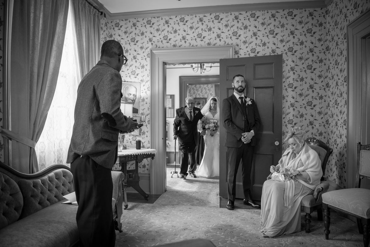 Bride and older man approach nervous groom by doorway in softly lit vintage room with camera adjuster and seated woman.