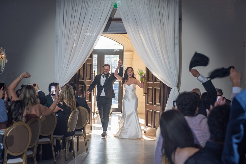 A smiling bride and groom run down the aisle, arms raised, as guests cheer and toss hats in celebration.