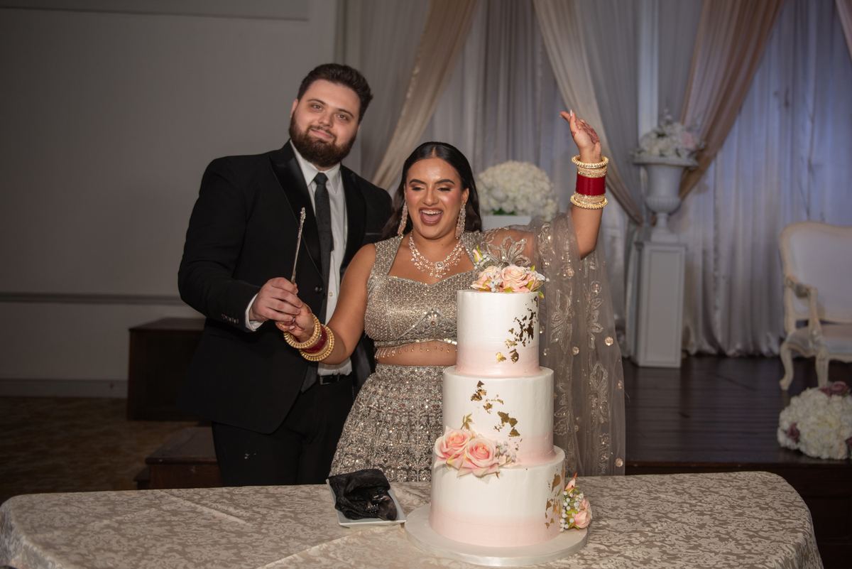 A bride and groom celebrate with a ceremonial knife beside their ornate wedding cake, adorned with floral and gold details.