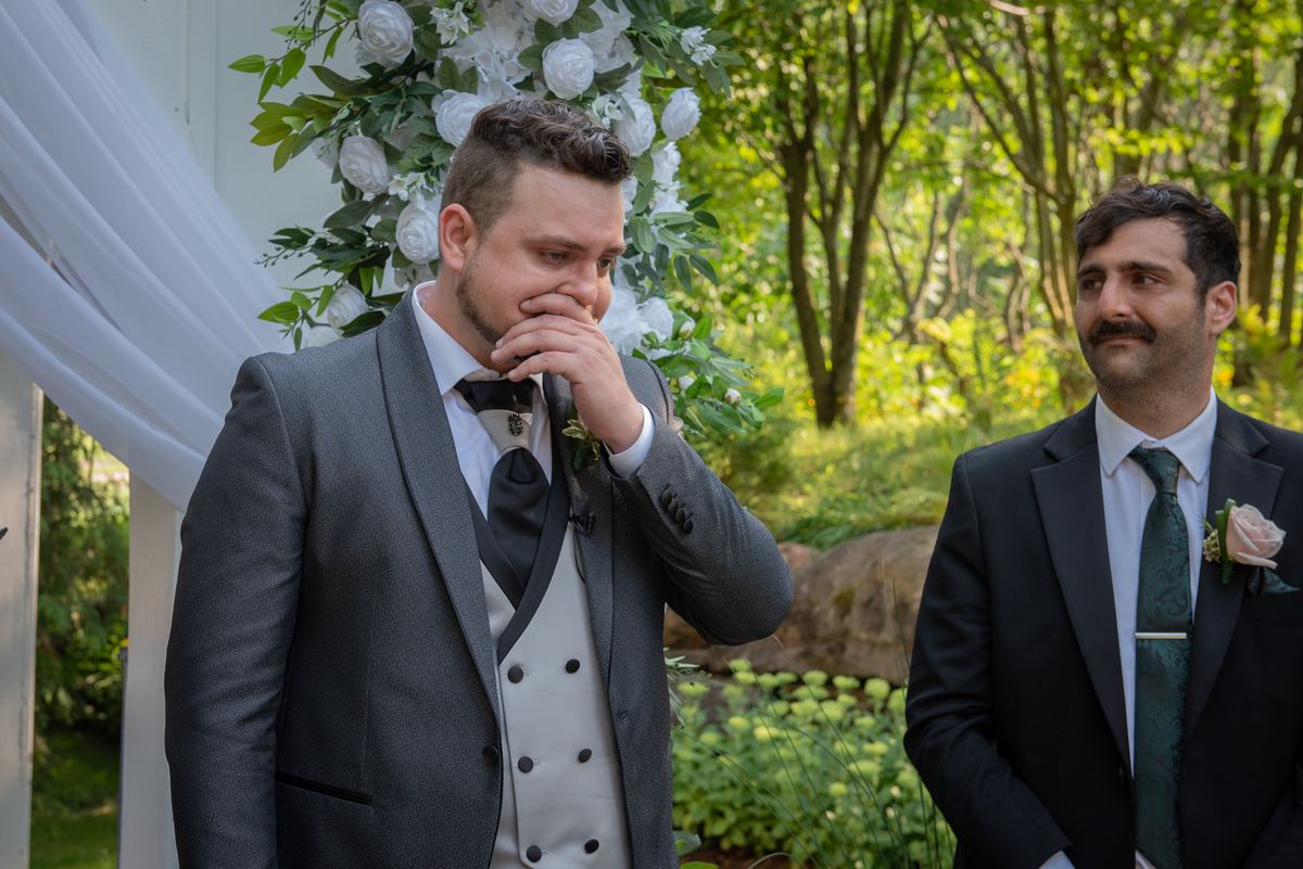A groom in a gray suit wipes his eye as he looks toward the ceremony, with a floral archway and his groomsman beside him.