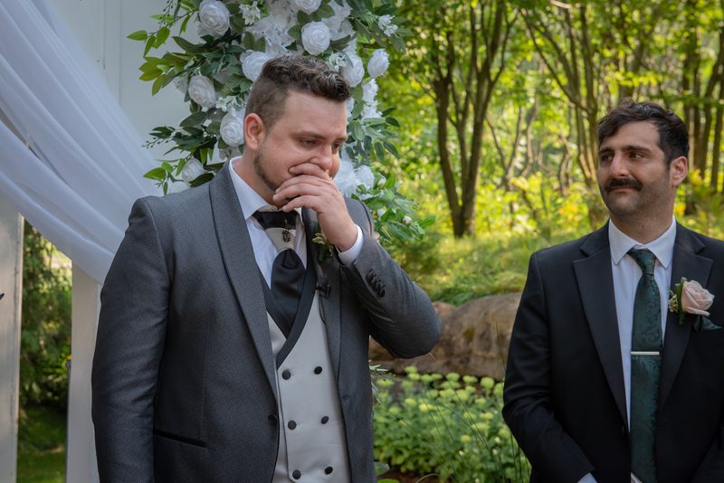 A groom in a gray suit wipes his eye as he looks toward the ceremony, with a floral archway and his groomsman beside him.